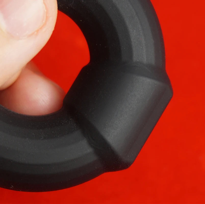 Black rubber constriction penis band held between fingers focusing in on the anchor buckle against a red background