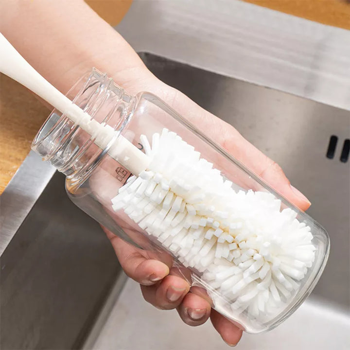 An image of a long white cleaning brush with a mop-like polyester head inside of a bottle cleaning it while someone holds the outside over a sink